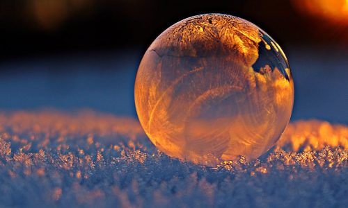 Close-up shot of a frozen bubble with warm reflections resting on a snowy surface at twilight.